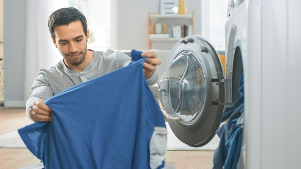 Handsome Smiling Young Man in Grey Jeans and Shirt Sits in Front of a Washing Machine at Home. He Loads the Washer with Dirty Laundry. Bright and Spacious Living Room with Modern Interior